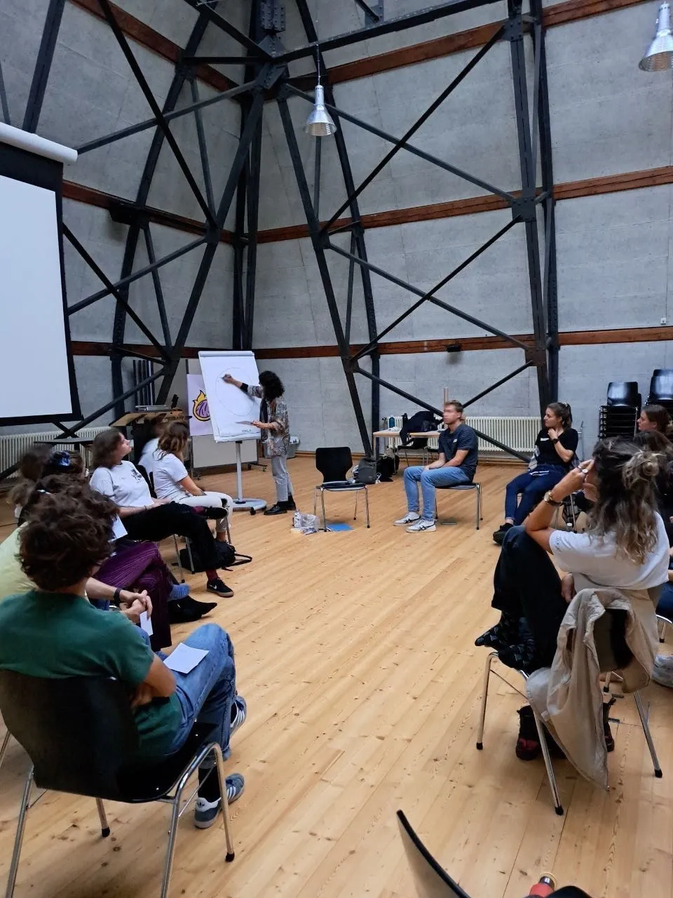 Menschen sitzen im Kreis, während eine Person am Flipchart etwas schreibt.