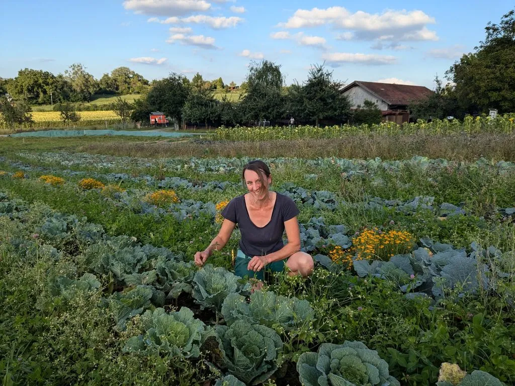 Junge Frau kniet mitten in grossem Gemüsebeet vor Bauernhof.