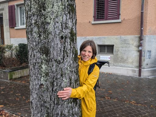 Lachende Frau in gelbem Mantel umarmt Baum.