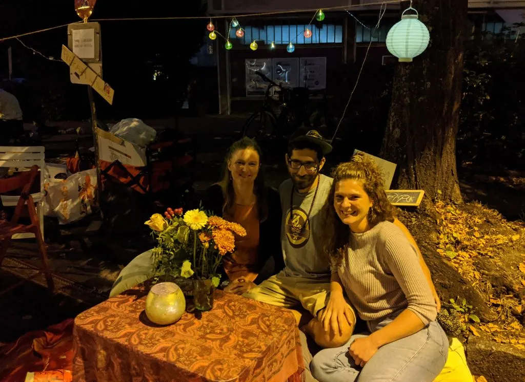 Drei junge Menschen sitzen auf Parkplatz mit Tisch und Kissen.